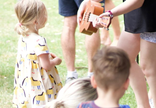 Music teacher holds an ukulele out to a young blonde girl